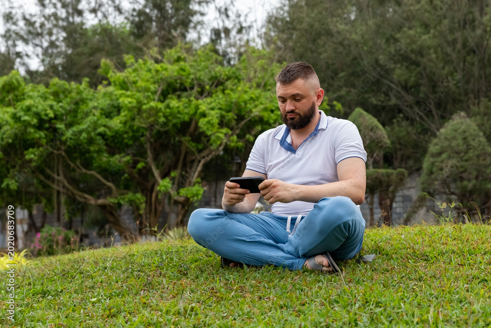 Obraz premium Man with a beard sitting cross-legged on green grass in a city park, holding a smartphone, with lush trees and shrubs providing a serene backdrop in an outdoor nature environment