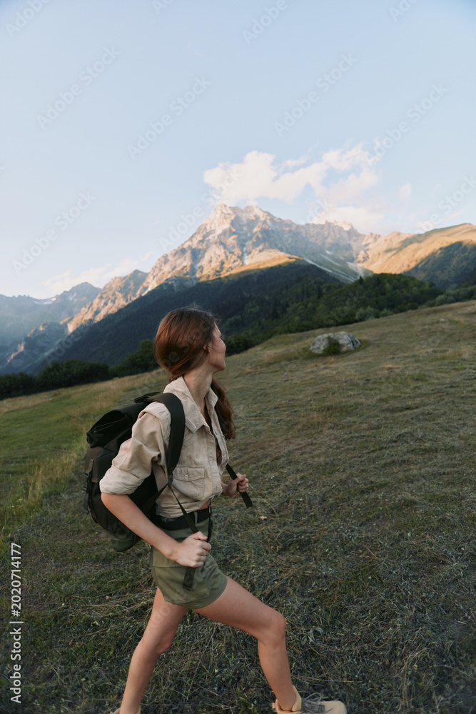 Obraz premium Hiking woman with a backpack strides through a grassy meadow toward distant mountains, focused and determined in a natural outdoor setting, her hair tied back, soft light highlighting the landscape