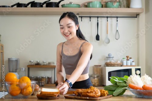 A smiling fit Asian woman in athletic wear prepares fresh ingredients on a wooden kitchen counter, ideal for health, wellness, and lifestyle content