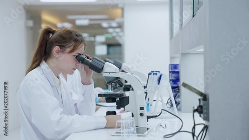 A woman looks closely at samples using a microscope in a bright laboratory. Another person works at a separate station. The area has lab equipment and supplies on the counters.