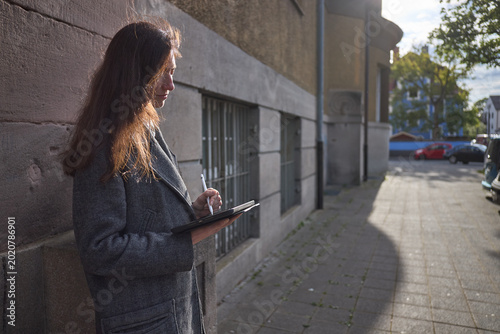 Woman sketching on a tablet device while leaning against a textured wall outdoors