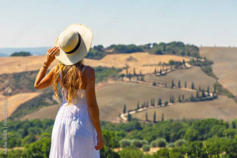 Obraz premium Back view of woman in white dress holding straw hat looking at cypress winding road in Val d'Orcia Tuscany. travel destination, summer vacation in Italy