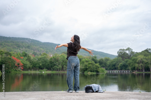 Foto Back view shot of asian woman opening both arms while standing beside the lake and mountain in park