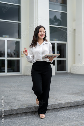 Full body of asian businesswoman holding tablet and pen while standing walking outside the building.
