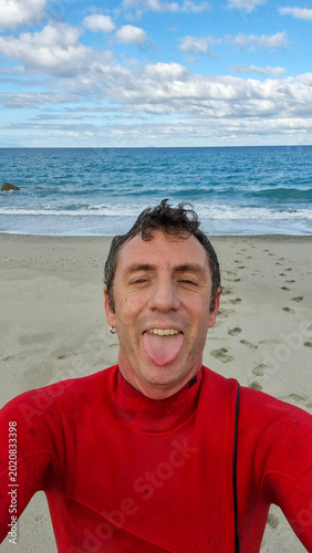 A man in a surf suit sticks his tongue out for happiness, behind him the sea horizon under a blue sky with white clouds. The photo was taken in the seaside village of Rodia, Sicily, in Southern Italy