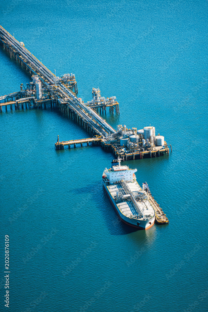Fototapeta premium A cargo ship is docked at an industrial port. The blue water surrounds the pier with many structures used for loading and unloading