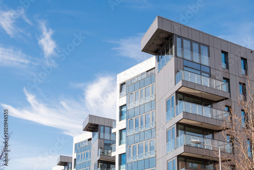 An upward angle shot of a modern multi-story apartment building with a sleek grey and white facade, glass balconies, and large windows. 