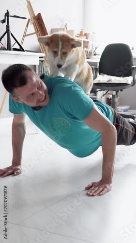 Young man doing push-ups at home while his playful Pembroke Welsh Corgi puppy stands on his back, enjoying fun exercise and companionship in a cozy domestic setting. 