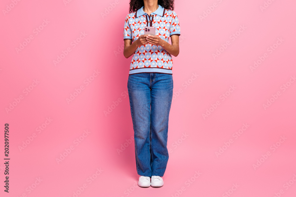 Naklejka premium Young woman in a patterned polo shirt jeans and white sneakers using a pink phone against a pink background for a stylish fashion lifestyle feel
