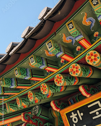 Traditional korean dancheong coloring on roof eaves. Close-up of wooden roof eaves with decorative coloring of vibrant green, red, and orange floral patterns under a clear blue sky. 