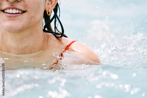 A smiling young woman with wet hair and a red swimsuit is enjoying a refreshing swim in the water. Water splashes around her as she relaxes and has fun.