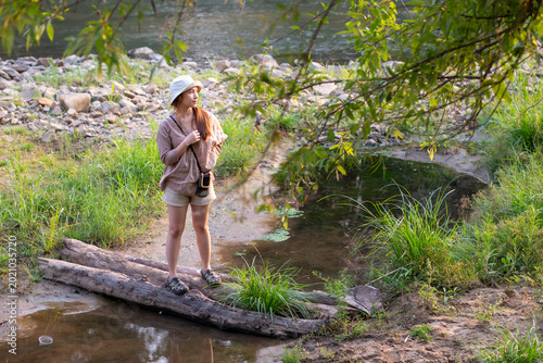 Fotografie Young woman wearing hat holding camera standing on log over stream looking peace