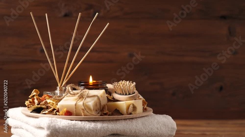 Still life of a home spa set on a wooden background. Burning candle in a glass jar, natural handmade soaps, aroma diffusers, and dried petals on a white towel. Calm and zen atmosphere for wellness.