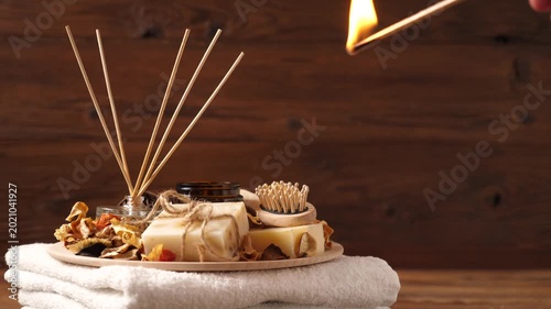 Close-up of a hand lighting a candle in a home spa set. Composition with natural soaps, essential oils, aroma sticks, and a white towel on a wooden background. Relaxation and wellness atmosphere.