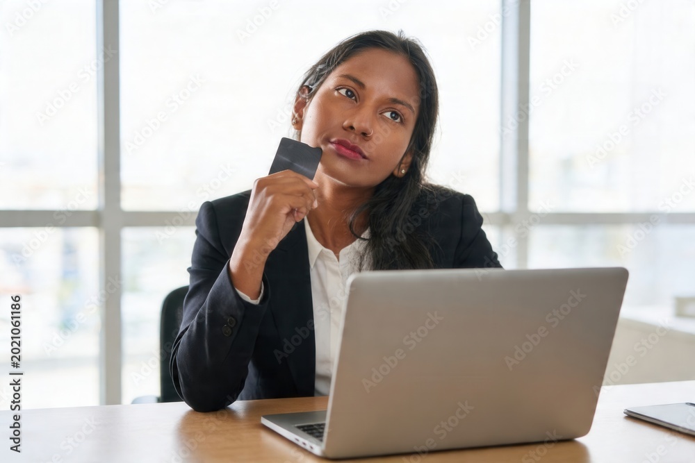 Fototapeta premium Young businesswoman contemplating an online purchase holding a credit card and using a laptop