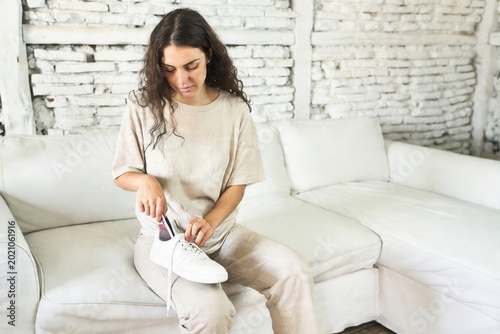 Woman sitting on white sofa inserting foot into white sneakers