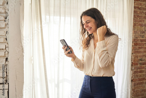 Woman standing by window looking at smartphone, celebrating success with a fist pump