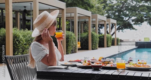 Woman drinking orange juice at dining table, outdoor luxury resort patio beside swimming pool with cabanas, summer vacation wellness, hotel breakfast service, upscale travel marketing