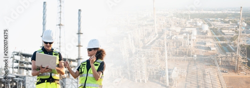 Construction workers conduct oil refinery inspection atop towering crane against sprawling industrial complex, utilizing tablets and radios for detailed maintenance assessments.