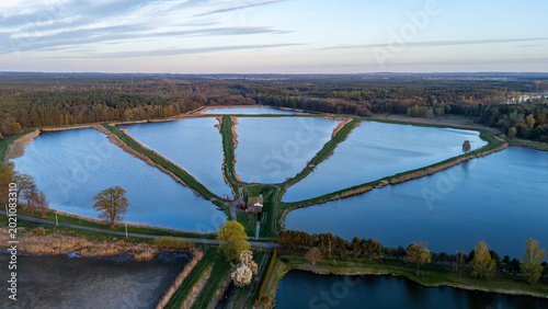 Cuadro en lienzo High-angle aerial view of a fish farm pond complex separated by narrow grassy dikes, surrounded by a vast forest and rural landscape during golden hour with calm water reflecting the soft sky