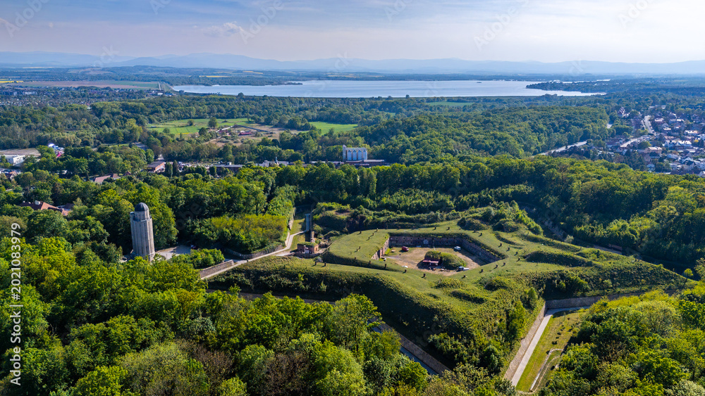 Obraz premium Panoramic aerial view of Fort Prussia in Nysa, showcasing geometric grass-covered bastion fortifications, a stone tower, the vast Nysa Lake, and a mountain range on the horizon in bright daylight.