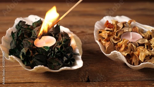 Close-up of a hand lighting tea light candles nestled in decorative shells filled with colorful dried flower petals. Cozy home spa decor on a wooden background, creating a relaxing atmosphere.