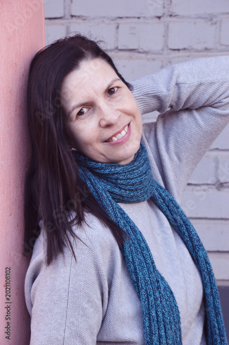 Slika na platnu A dark-haired Caucasian woman wearing a turquoise scarf stands, pressing her hand to her neck and looking at the camera