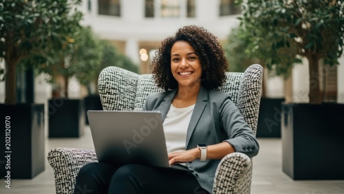 Smiling woman using laptop, seated in elegant chair, in atrium