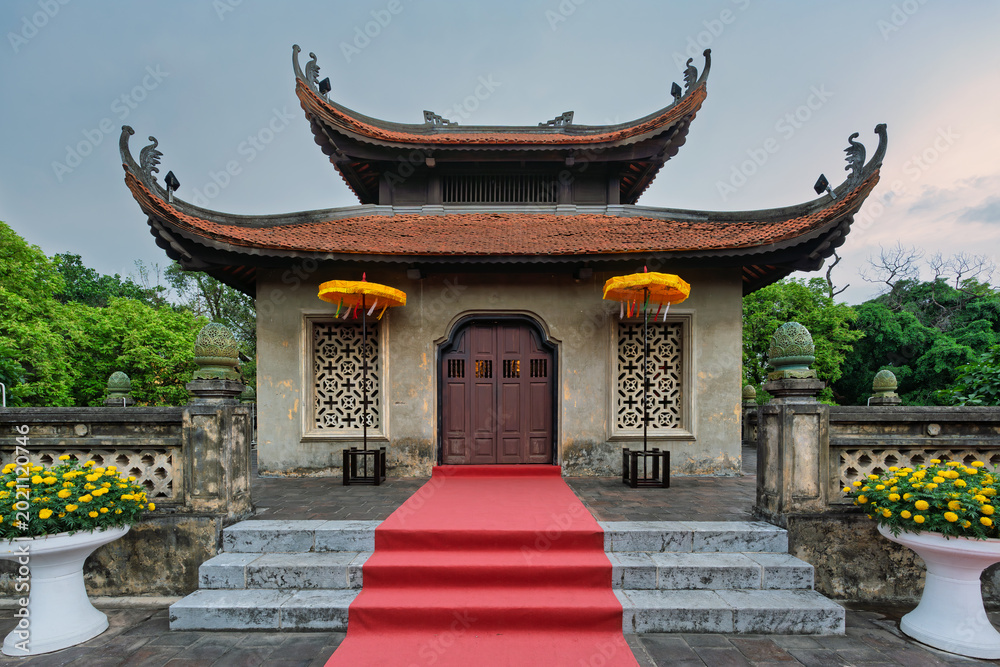 Fototapeta premium Traditional Vietnamese architecture of North Gate Cua Bac, Imperial Citadel of Thang Long, Hanoi, Vietnam. Featuring red tiled roof, ornate details, a welcoming red carpet, and vibrant yellow flowers