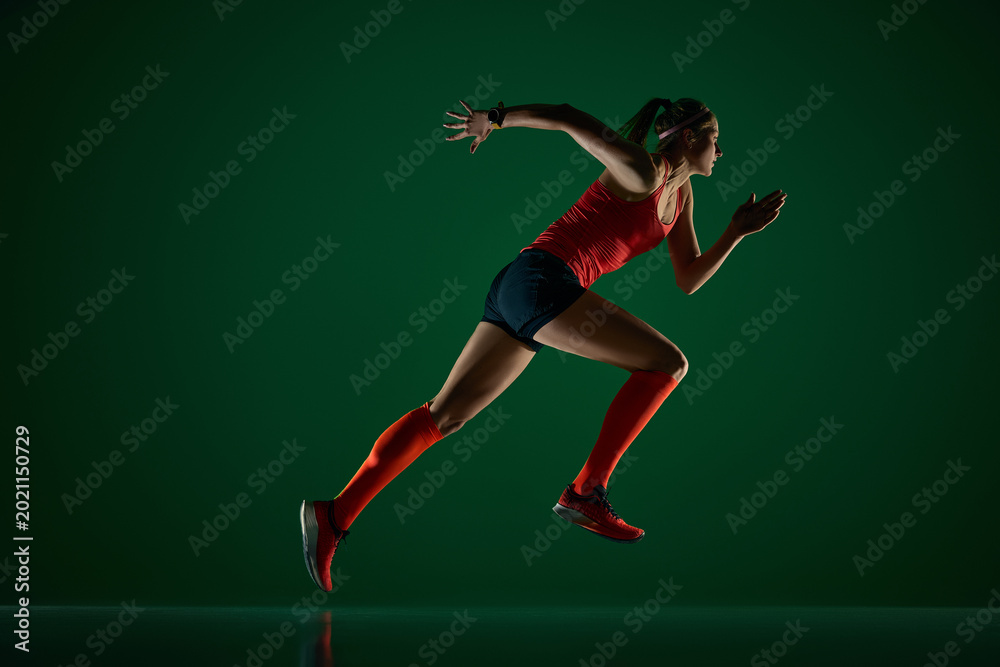 Fototapeta premium Focused woman runner sprinting forward on dark green studio floor. Concept of speed workout, sports performance, running apparel, fitness ads and active lifestyle branding.