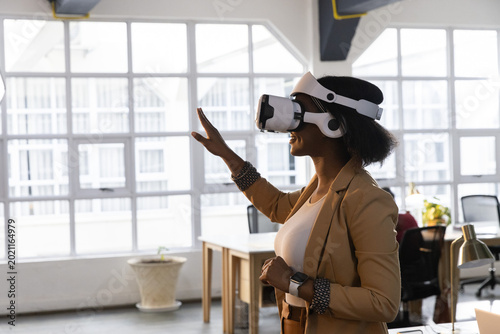 Obraz na plátne African American woman wearing VR, tan blazer reaching interface near office win