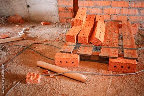 Schilderij op canvas A few unused red clay bricks lie on a makeshift wooden pallet alongside construction debris and a loose wire on the dusty floor of an unfinished room