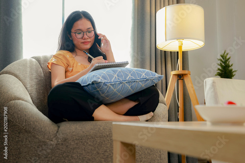 The lifestyle Asian woman: sitting calmly on a sofa using her smartphone she seriously jotted down phone numbers and messages in a notebook.
