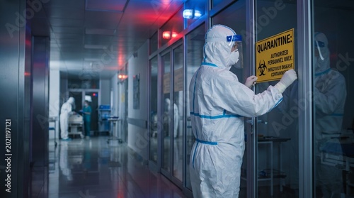 Healthcare worker in a hazmat suit and face shield, affixing a quarantine sign to a hospital door. Urgent infectious disease containment in a sterile corridor.