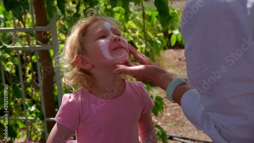 Close up of a mother hands applying white sunscreen cream on her little daughter face to protect skin from UV rays. Family enjoying a sunny day in a green garden