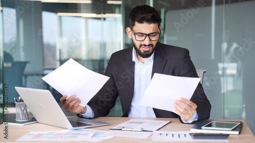 Confused puzzled businessman having difficulty with paperwork sitting at desk in business office. Frustrated manager unhappy with bad financial results reviewing documents and looking at laptop screen