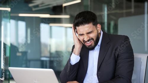 Unhealthy businessman in suit suffers from ear pain sitting in office. Sick male touches his ear with his fingers. He has furuncle, shingles, eczema, ear diseases or otitis media. Earache. Close up