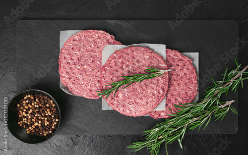 Raw beef burger patties on a black slate board with rosemary and peppercorns.