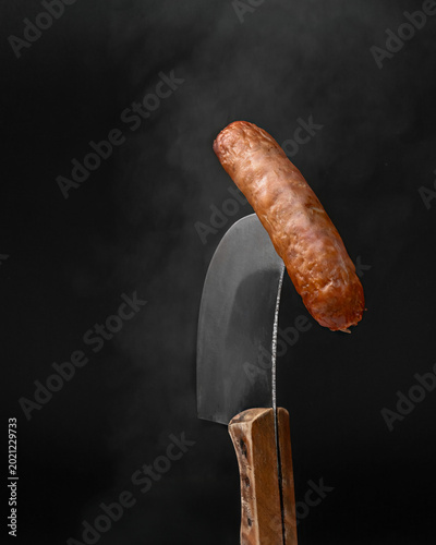 Grilled juicy sausage balanced on a sharp meat cleaver with steam on black background.