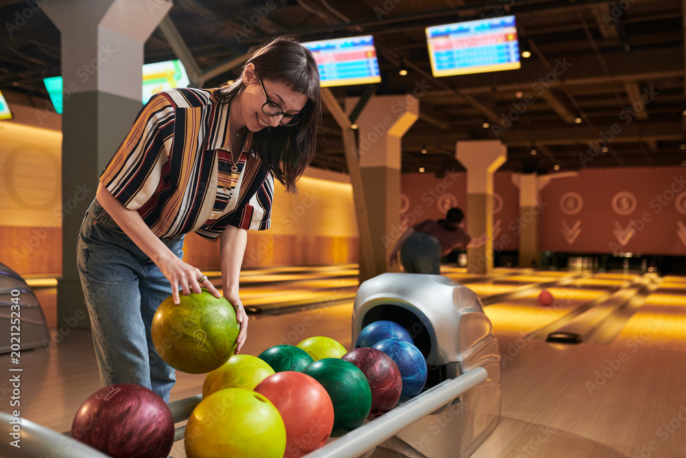 Fototapeta premium Young adult woman selecting bowling ball at alley, preparing game and leisure activity. Suitable for entertainment marketing, team bonding promotion, lifestyle advertising