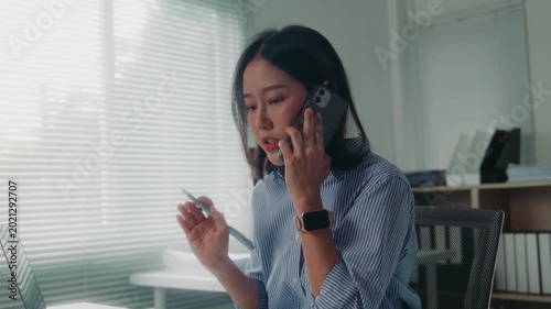 Anxious asian businesswoman feeling stressed and frustrated while talking on her mobile phone and working on her laptop at the office