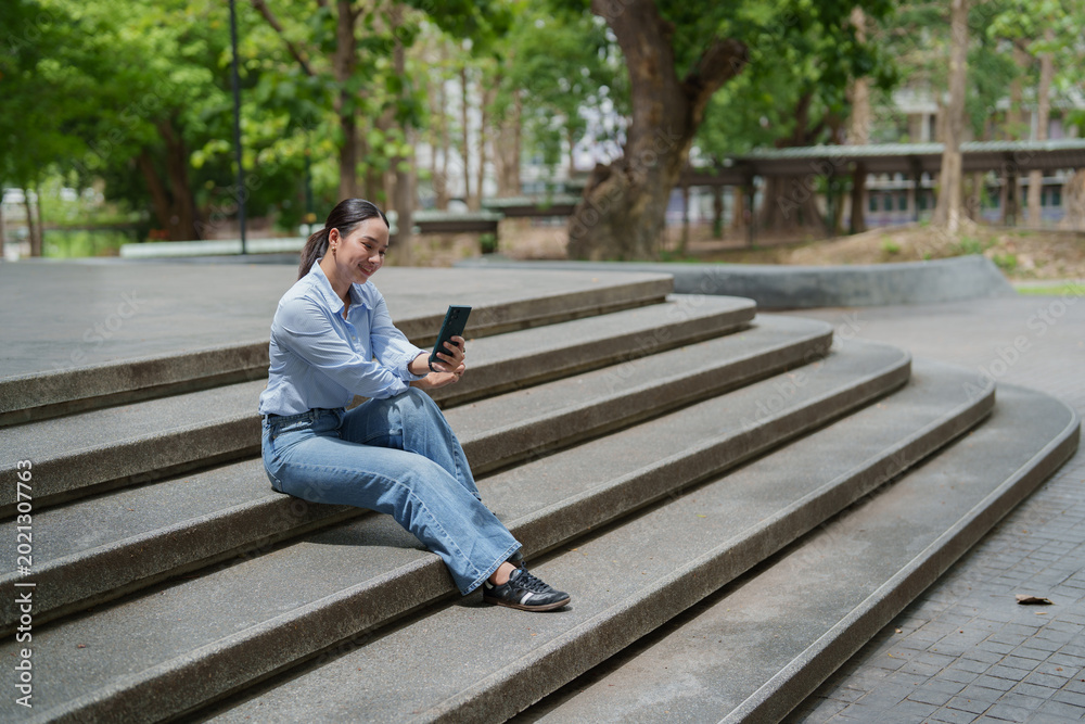 Fototapeta premium Woman sitting on campus steps using a mobile phone, engaging with digital content, representing modern communication, remote work, and online education