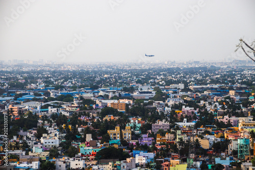A wide aerial view of a densely populated cityscape of chennai city in India with colorful buildings, greenery, and an airplane flying in the hazy sky.
