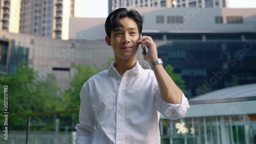 Confident young Asian businessman making a phone call outside modern office buildings. Corporate communication and professional business lifestyle in urban environment.
