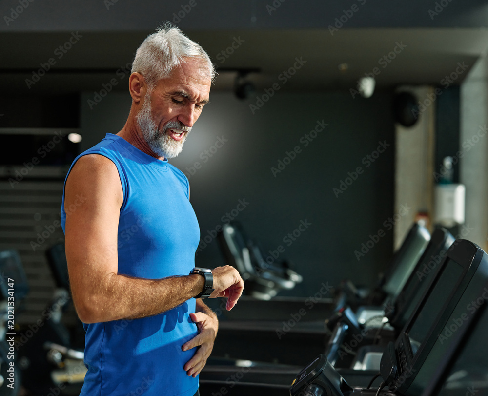 Obraz premium Portrait of a senior man exercising in a gym, mature male running using treadmill machine and checking smart watch for performance, healthy lifestyle and cardio exercise at fitness club concepts, vita