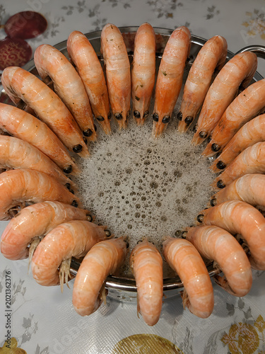 Fresh boiled shrimp arranged in a circular composition on a metal tray with foamy seafood broth in the center, close-up food presentation.