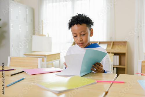 Obraz na plátně A young boy attentively reads a workbook in a bright classroom with colorful stationery scattered on desks