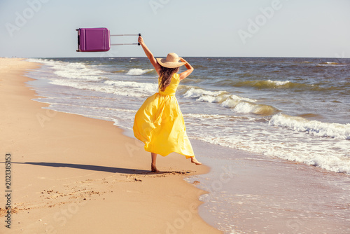 Young happy woman in a yellow long dress with hands in the air runs on the seaside in summer. The woman holds travel suitcase. Summertime, travel, and happy holiday concept