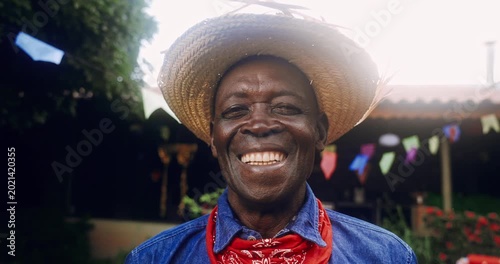 A joyful elderly Black man wearing a traditional straw hat and a red bandana smiles warmly at the camera during a festive outdoor celebration with colorful flags in the background