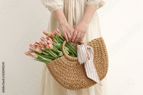 Woman holding wicker bag with beautiful tulips and handkerchief on light back...